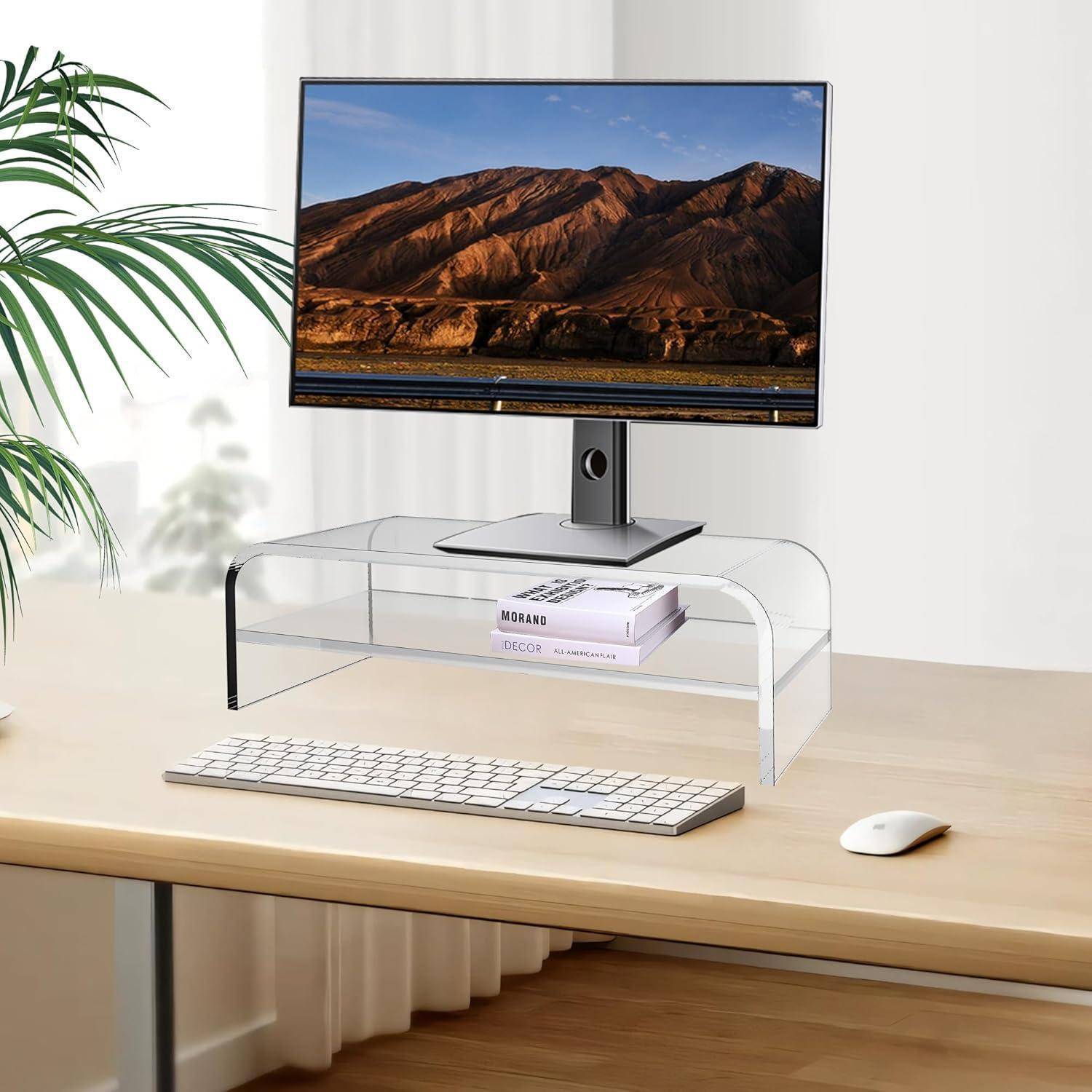 Ein minimalistischer Schreibtisch mit einem Monitor, der eine Berglandschaft zeigt. Darunter hält ein Laptopständer ein Buch und ein Telefon. Eine Pflanze links daneben.