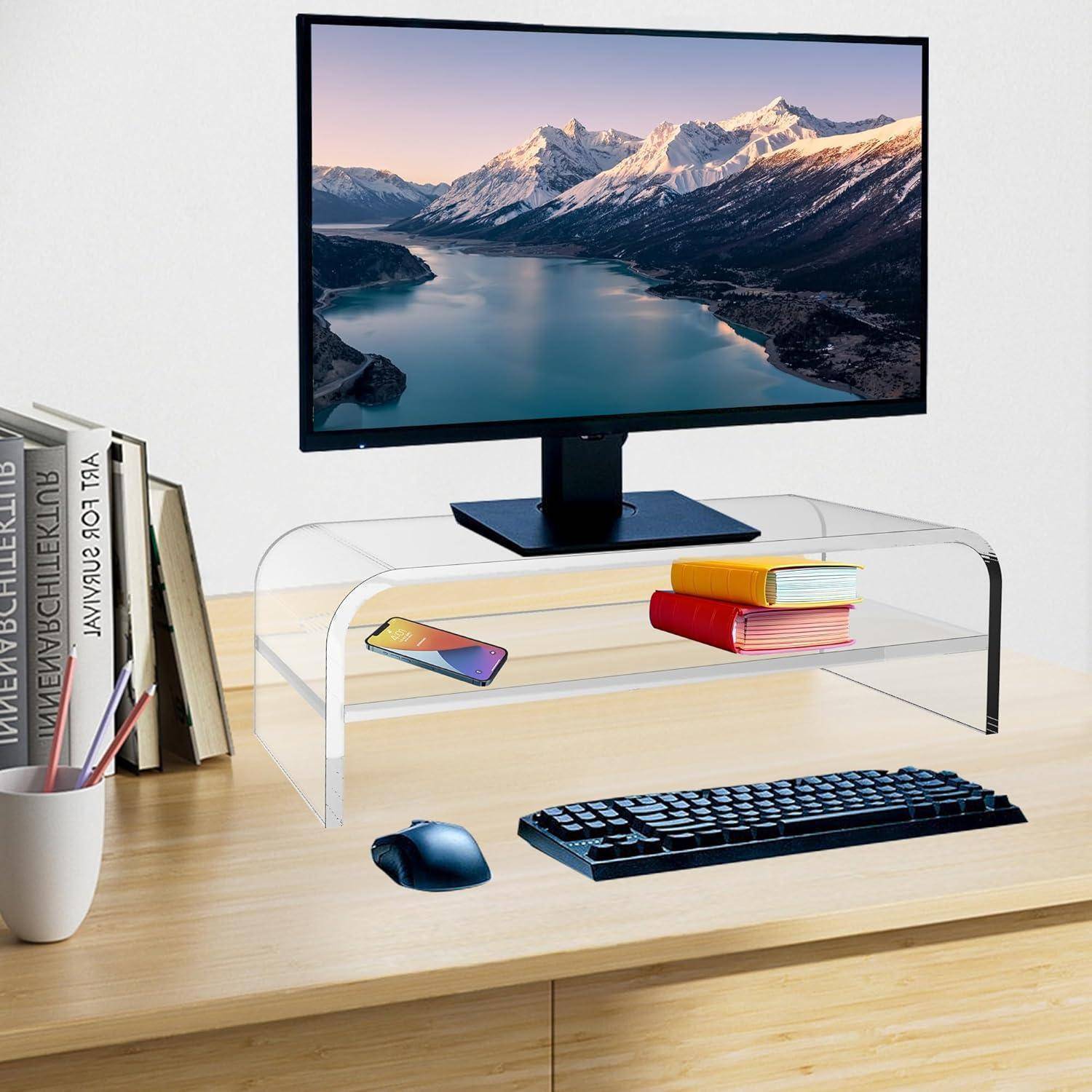 Ein Monitor, der eine Berglandschaft zeigt, steht auf einem Schreibtisch mit einem transparenten Ständer. Darunter sind Tastatur, Maus, Telefon und Bücher ordentlich angeordnet.