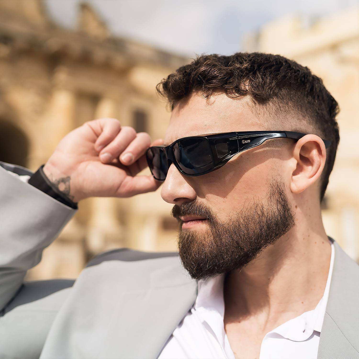 Eine Person mit schwarzer Sonnenbrille und einem hellgrauen Anzug steht im Freien, wobei historische Architektur im Hintergrund unscharf ist.