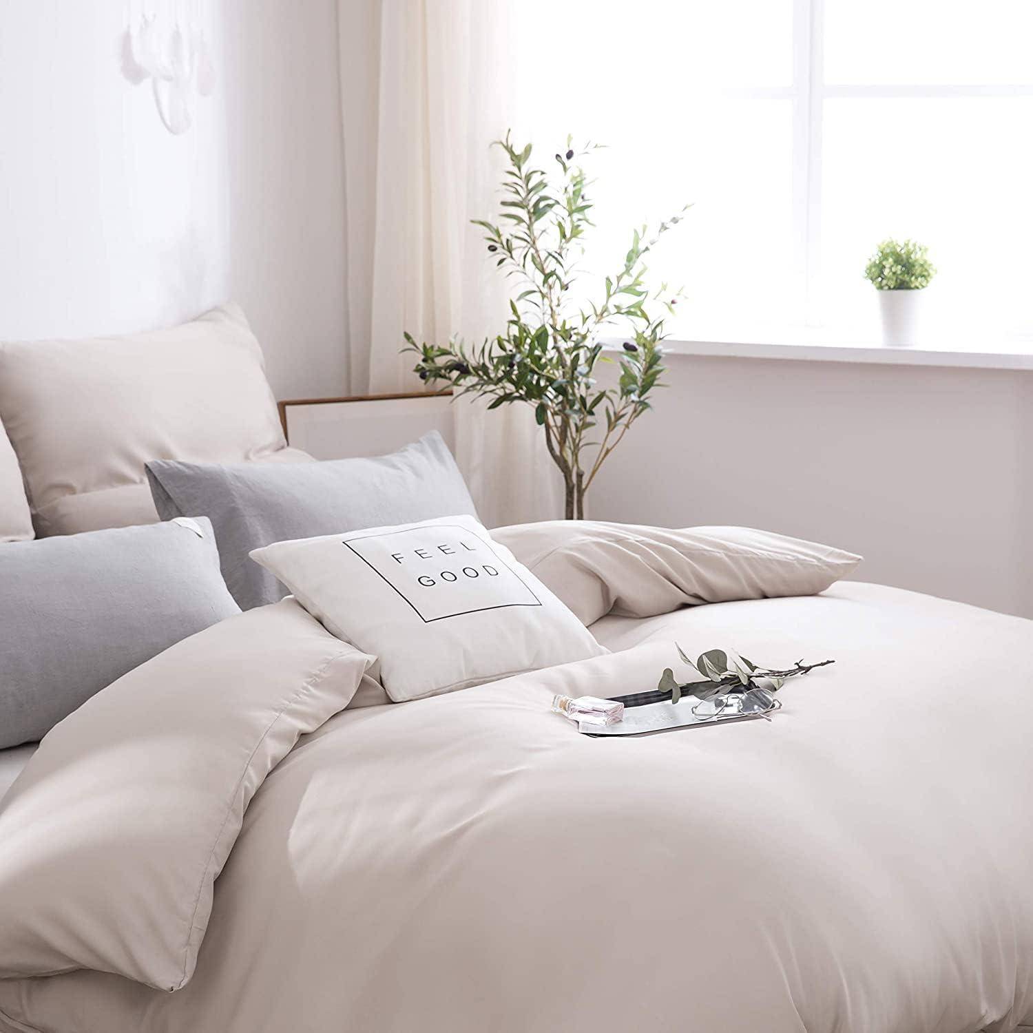 Ein sauber gemachtes Bett mit weißen und beigen Kissen und einer Bettdecke. Eine kleine Pflanze steht auf einem Nachttisch. Natürliches Licht von einem Fenster erhellt den Raum.