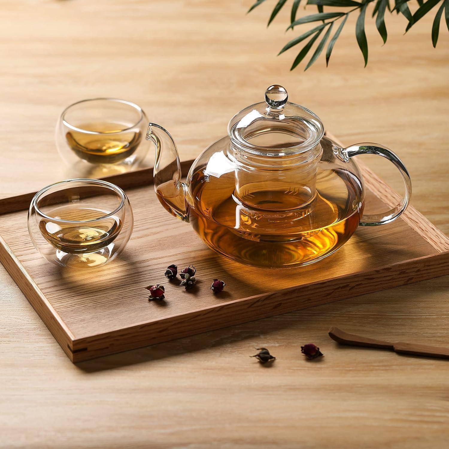 Eine Glasteekanne gefüllt mit Tee und zwei Teebechern auf einem Holztablett, mit verstreuten losen Teablättern und einem Zweig darüber.