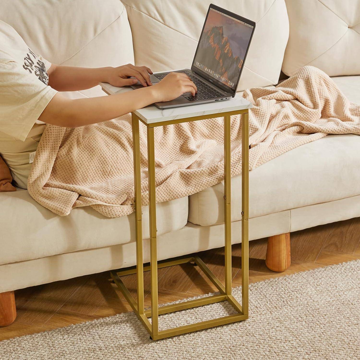 Eine Person sitzt auf einem Sofa, arbeitet an einem Laptop auf einem kleinen Ständer und ist in eine beige Decke gehüllt. Der Raum atmet eine gemütliche und entspannte Atmosphäre.