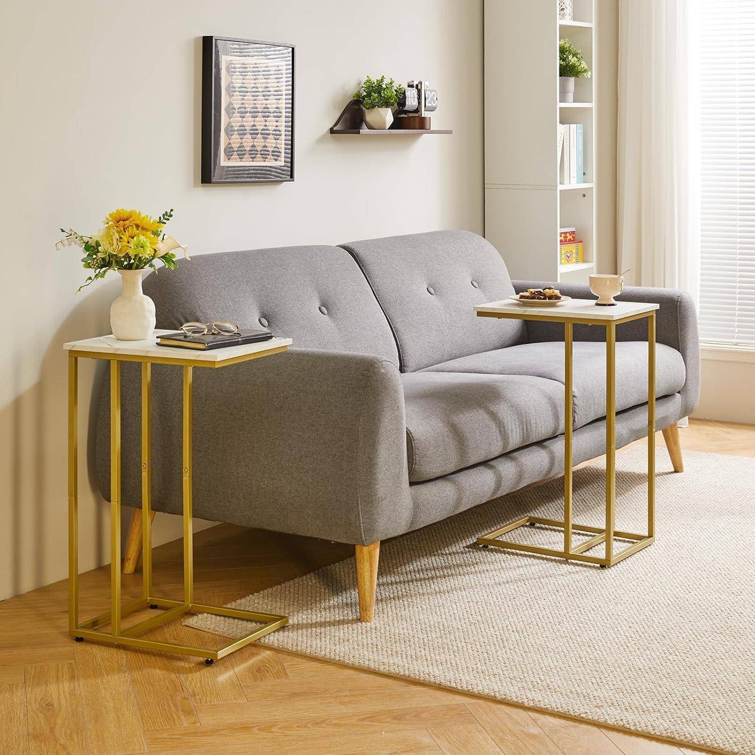 Ein moderner Wohnraum zeigt ein graues Sofa, das von zwei kleinen Tischen mit Pflanzen und Büchern flankiert wird. Natürliches Licht strömt durch ein Fenster auf der rechten Seite.