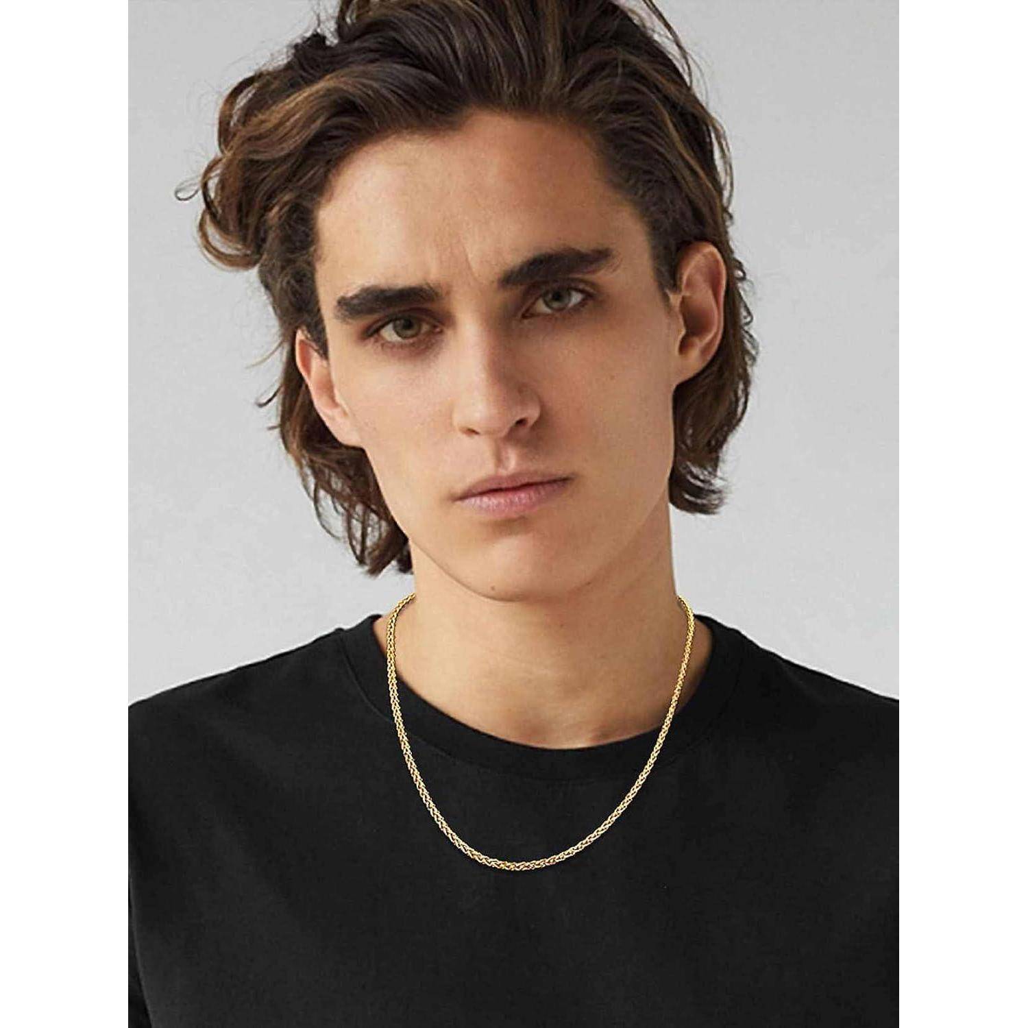 Eine Person mit langen dunklen Haaren, die ein schwarzes T-Shirt und eine goldene Halskette trägt und direkt in die Kamera blickt, vor einem neutralen Hintergrund.