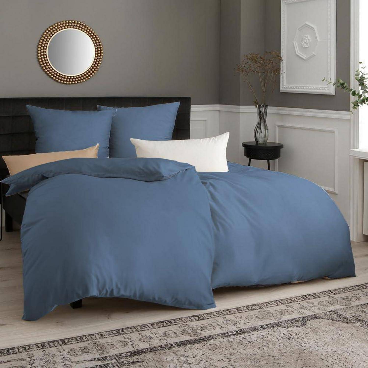 Ein ordentlich gemachtes Bett mit blauer Bettwäsche, zwei Kissen und einem weißen Dekokissen. Ein runder Spiegel hängt über einem dunklen Kopfteil. Eine Vase mit Zweigen steht auf einem Nachttisch.