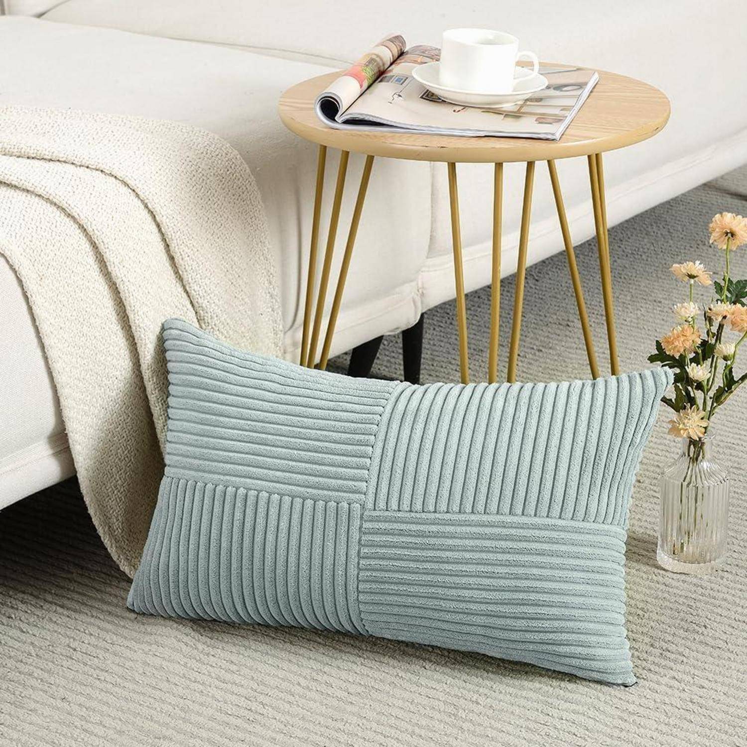 Ein hellblauer gerippter Kissen lehnt an einem beigen Sofa, und daneben steht ein runder Holztisch, auf dem eine Teetasse, Untertasse und ein Buch platziert sind.