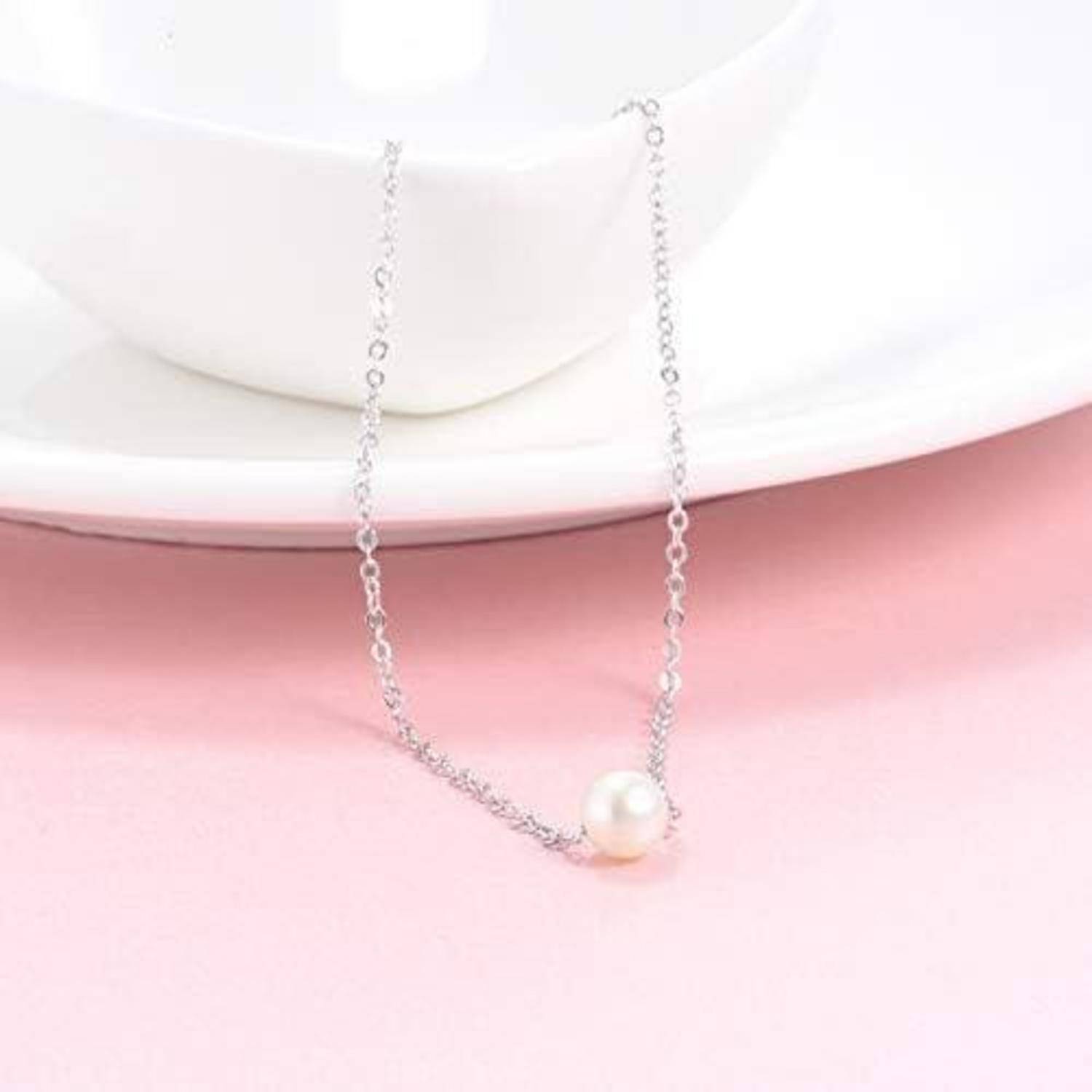 Eine Silberkette mit einem einzelnen Perlenanhänger liegt über einer weißen Schale vor einem rosa Hintergrund.