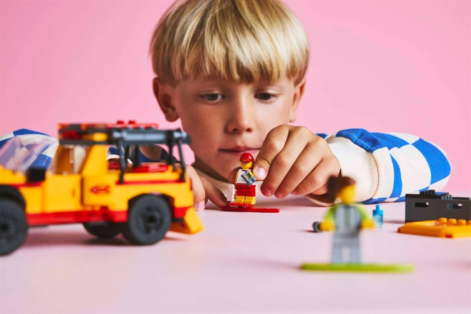 Ein junger Junge spielt mit LEGO-Figuren und einem Spielzeugfahrzeug auf einer rosa Tischplatte und konzentriert sich darauf, die Teile anzuordnen.