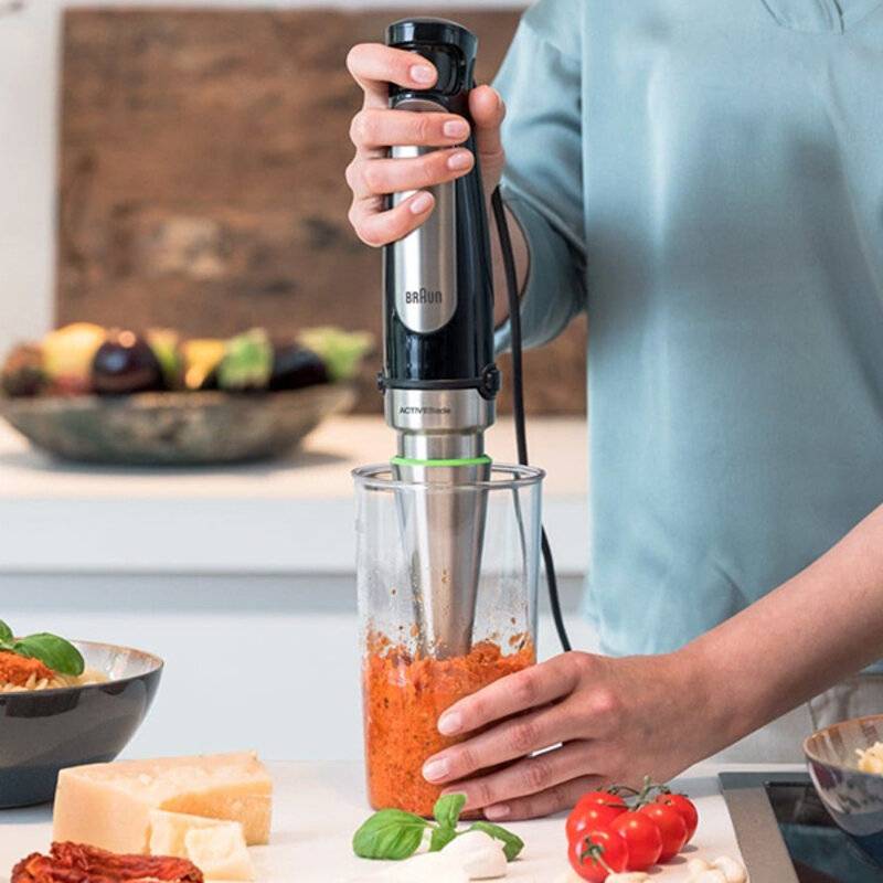 Eine Person, die einen Stabmixer verwendet, um eine orangefarbene Sauce in einem durchsichtigen Behälter zu mischen, wobei gehackte Gemüsestücke auf einer Küchenarbeitsplatte liegen.