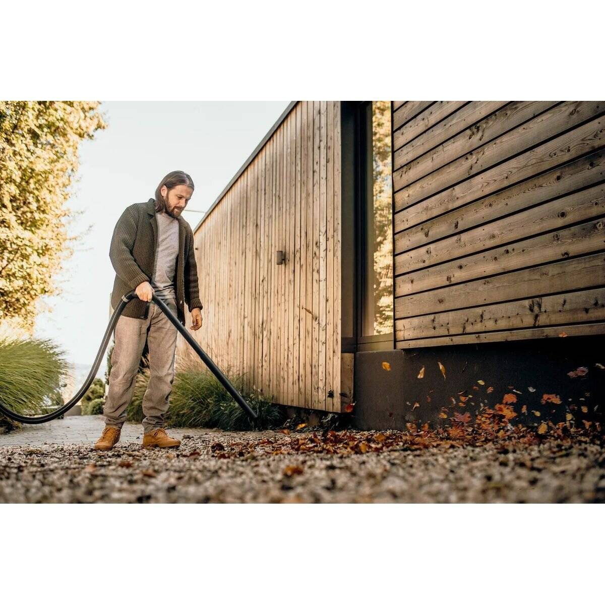 Eine Person harkt Herbstblätter von einem Kiesweg neben einem modernen Holzgebäude. Üppiges Grün umgibt die Szene und deutet auf einen Gartenbereich hin.