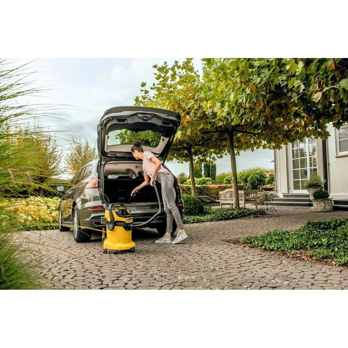 Eine Person platziert einen Staubsauger im Kofferraum eines Autos, das auf einer Kopfsteinpflasterdurchfahrt geparkt ist, mit Bäumen und einem Haus im Hintergrund.