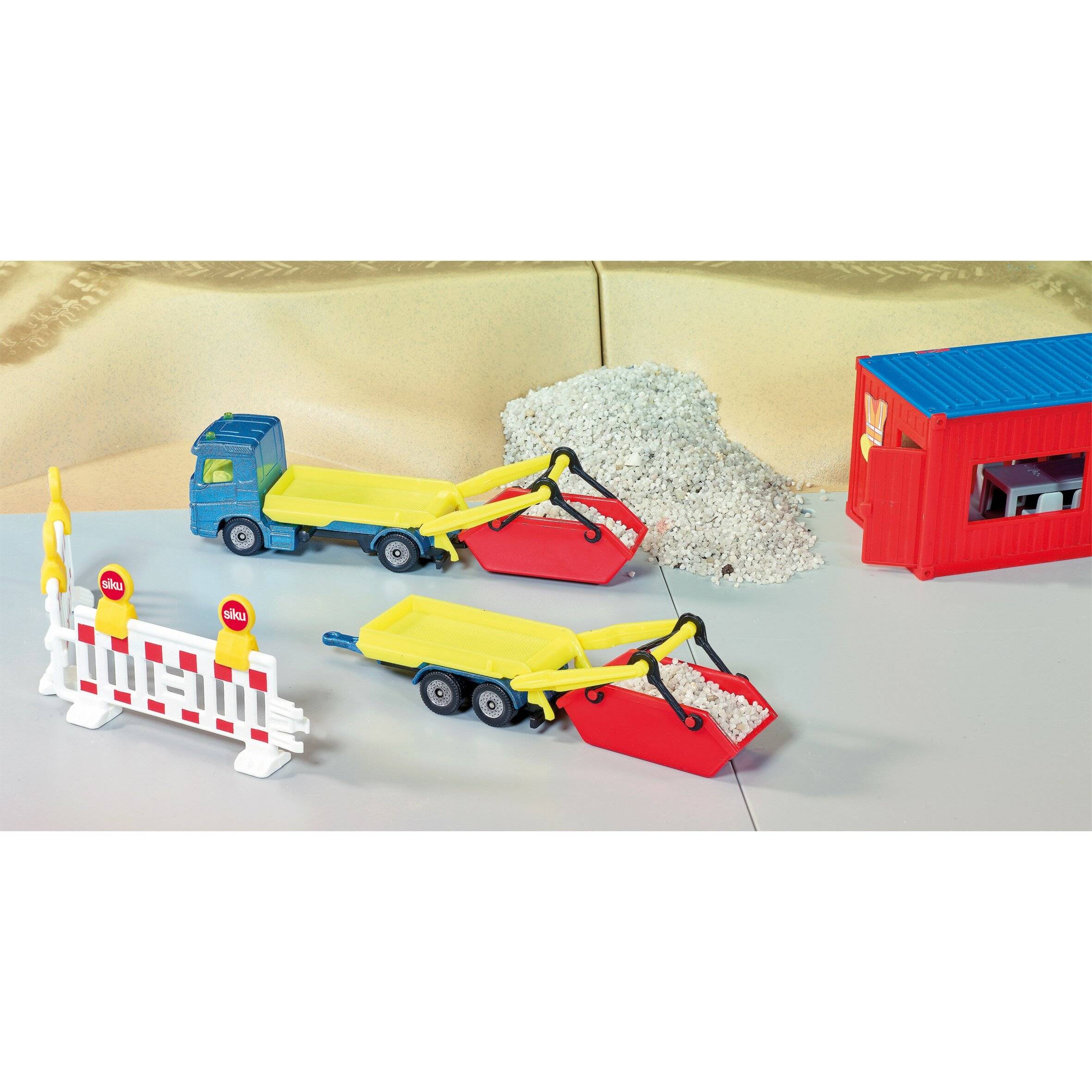 Spielzeug-Baustelle mit Spielzeug-Lastwagen, zwei Sandladern gefüllt mit Sand, einem roten Gebäude und Straßenabsperrungen auf einem Tisch.