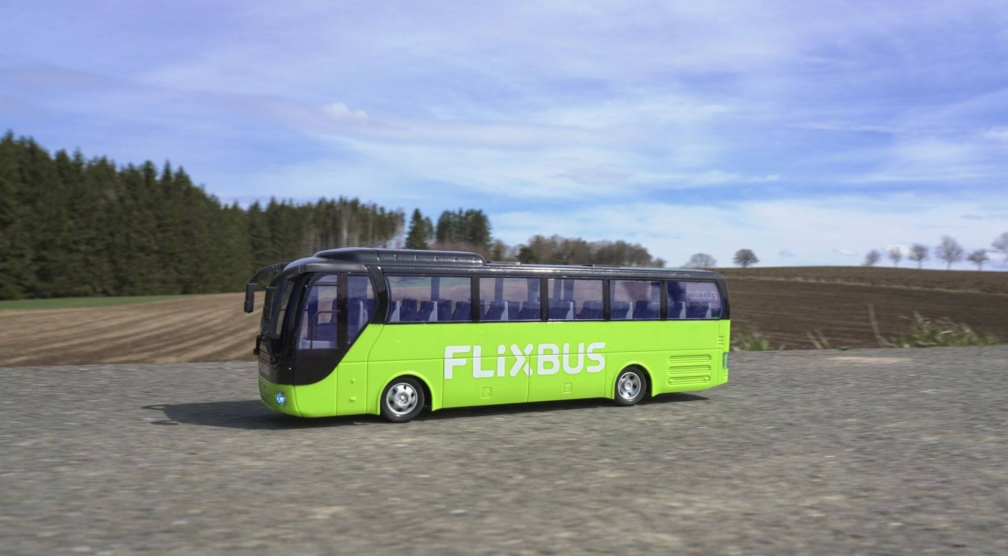 Ein grüner Bus mit dem 'FlixBus'-Logo fährt auf einer Landstraße, umgeben von Feldern und Bäumen unter einem teilweise bewölkten Himmel.