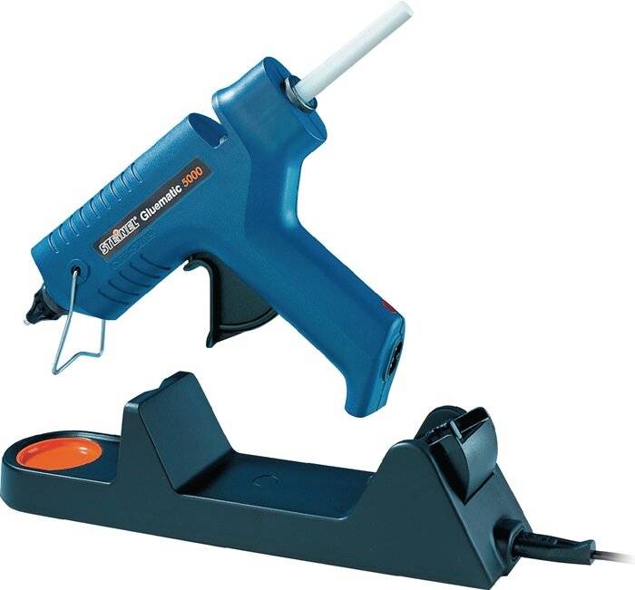 Eine blaue Heißklebepistole mit der Aufschrift "Glumatic 5000"", in die ein weißer Klebestift eingesetzt ist, die auf einem schwarzen Ständer ruht, vor einem einfarbigen weißen Hintergrund.