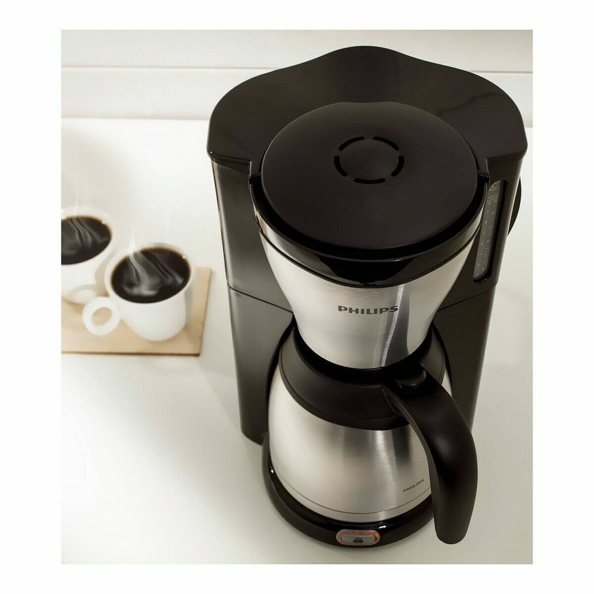 Ein silber-schwarzer Kaffeemaschine mit einer Glaskanne steht auf einer Arbeitsplatte. Zwei weiße Tassen, gefüllt mit schwarzem Kaffee, liegen daneben auf einer Serviette.