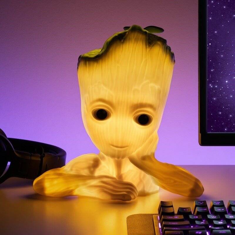 Eine kleine Gestalt, die einem Baum ähnelt, mit leuchtenden Merkmalen, sitzt auf einem Schreibtisch neben einem Computermonitor in einem dämmrigen Raum.