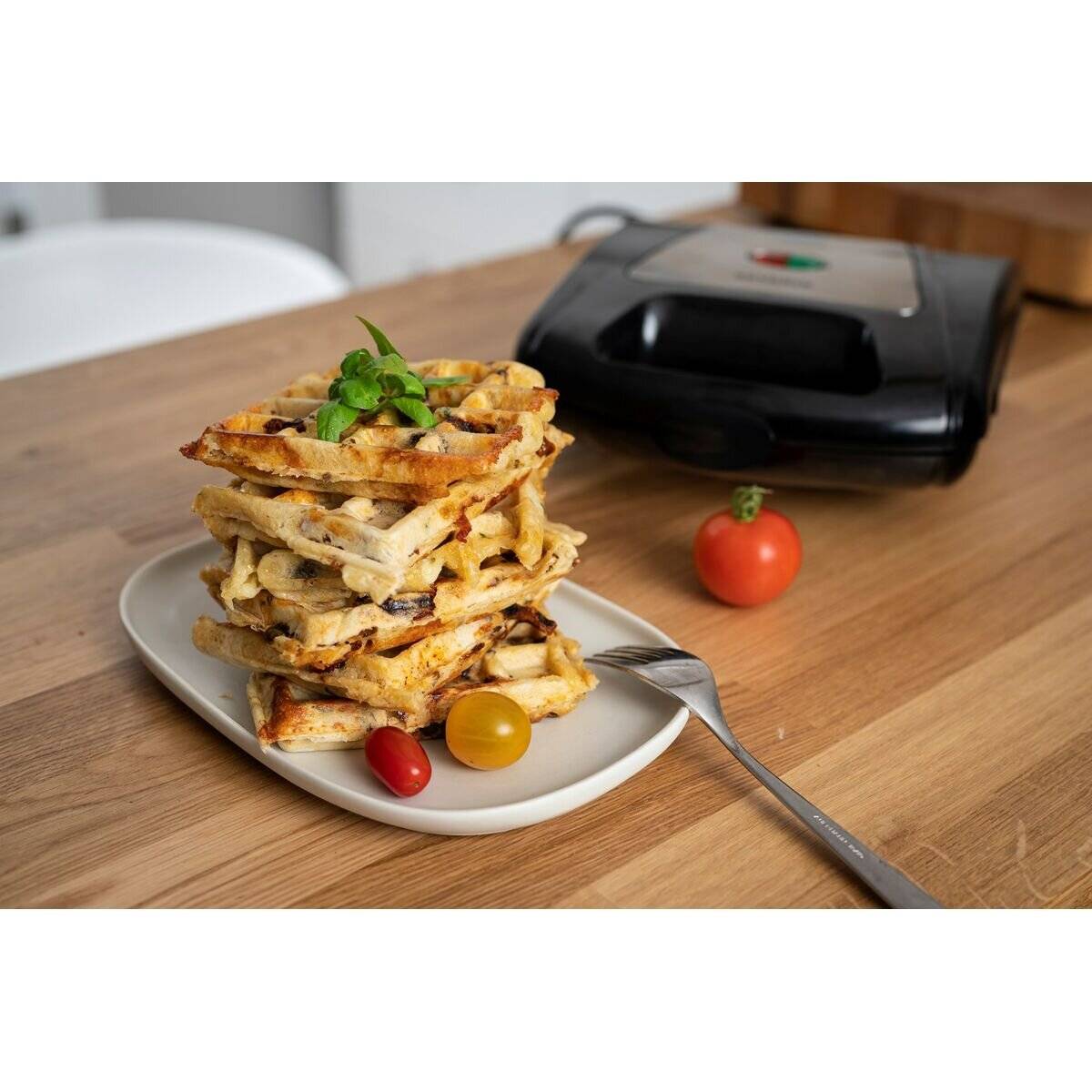 Ein Stapel Waffeln, garniert mit Minzblättern auf einem Teller, begleitet von Cocktailtomaten und einer Gabel, mit einem Waffeleisen im Hintergrund.