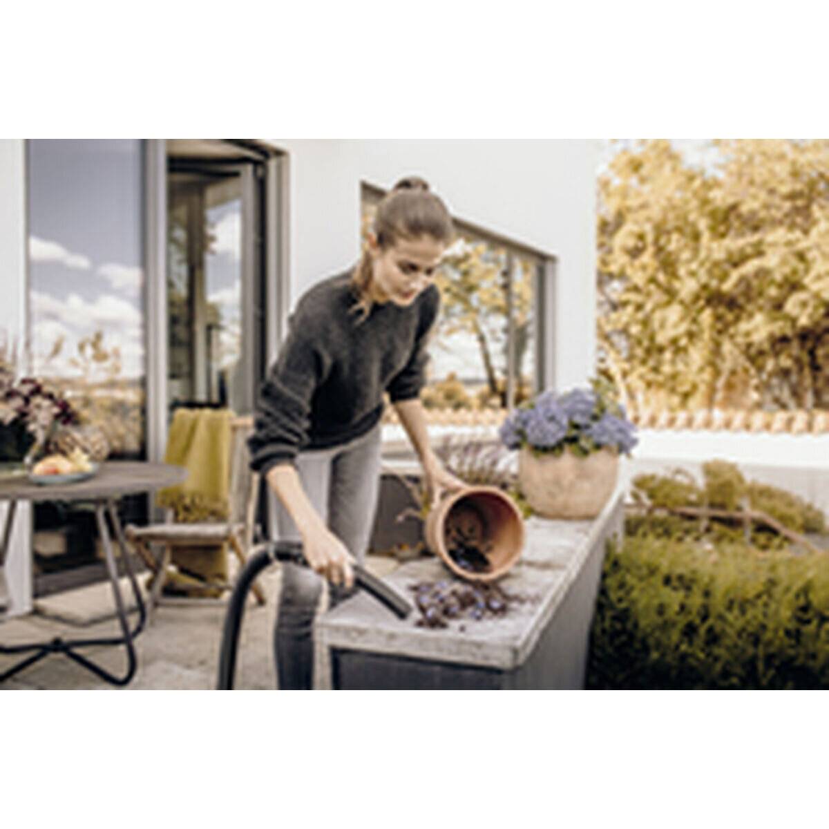 Eine Frau in einem Garten reinigt eine Arbeitsplatte mit einem kleinen Staubsauger, umgeben von Blumen und Gartenmöbeln, unter einem sonnigen Himmel.