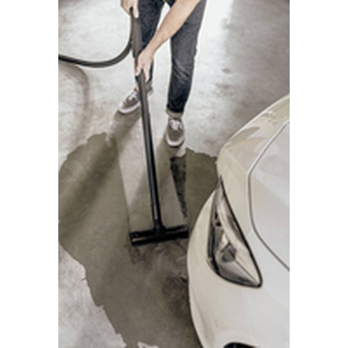 Eine Person in Jeans, die Wasser von einem Betonboden in der Nähe eines weißen Autos absaugt und wahrscheinlich ein Leck oder einen Wasserschaden in einer Garagenumgebung behebt.