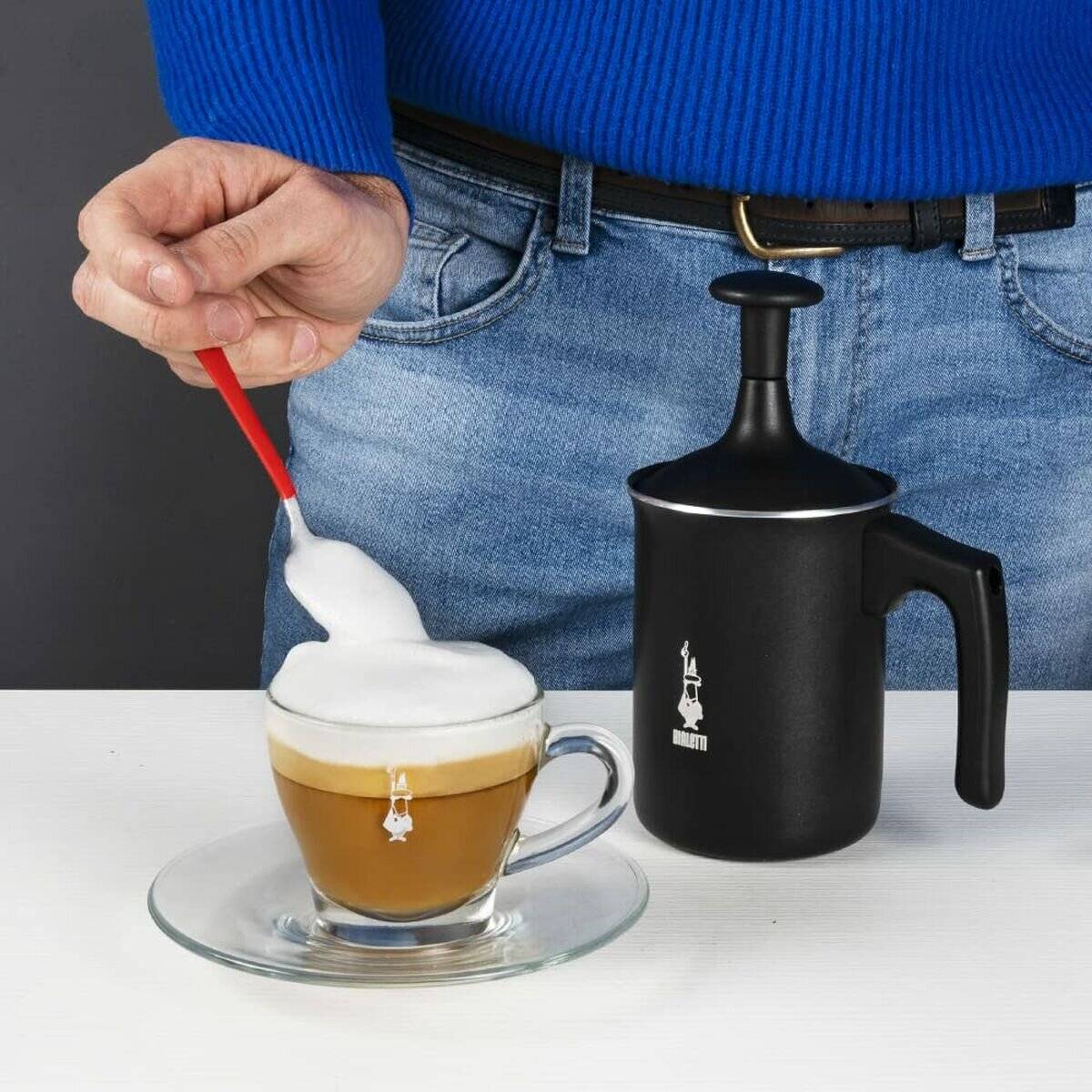 Eine Person, die einen roten Löffel mit aufgeschäumter Milch über einem Cappuccino in einer durchsichtigen Glasstasse hält, wobei ein Milchaufschäumer auf dem Tisch in der Nähe steht.