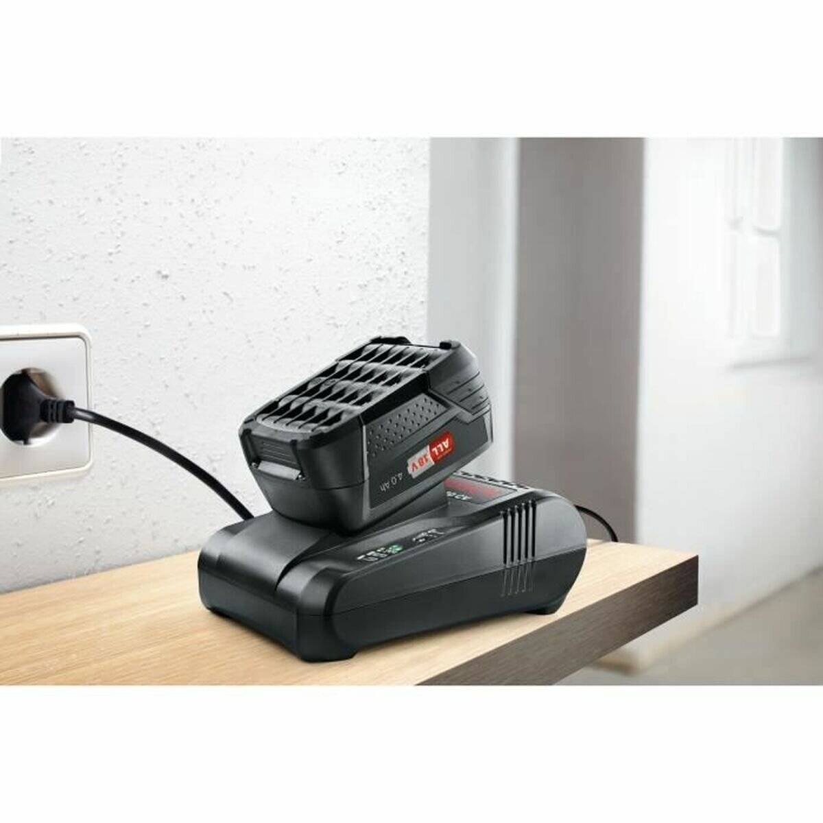 Eine schwarze Batterie lädt auf einem Holztisch, in eine Steckdose eingesteckt. Eine weiße Wand und ein Fenster sind im Hintergrund sichtbar.