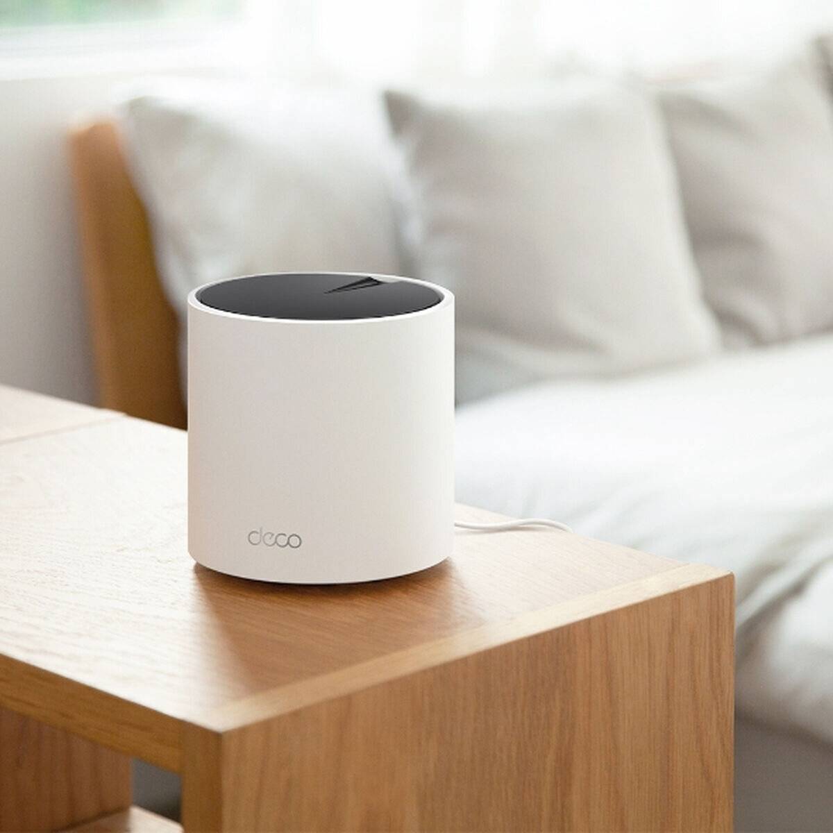 Ein weißes zylindrisches Smart-Home-Gerät mit schwarzem Deckel steht auf einem Holztisch in einer minimalistischen Wohnzimmeratmosphäre.