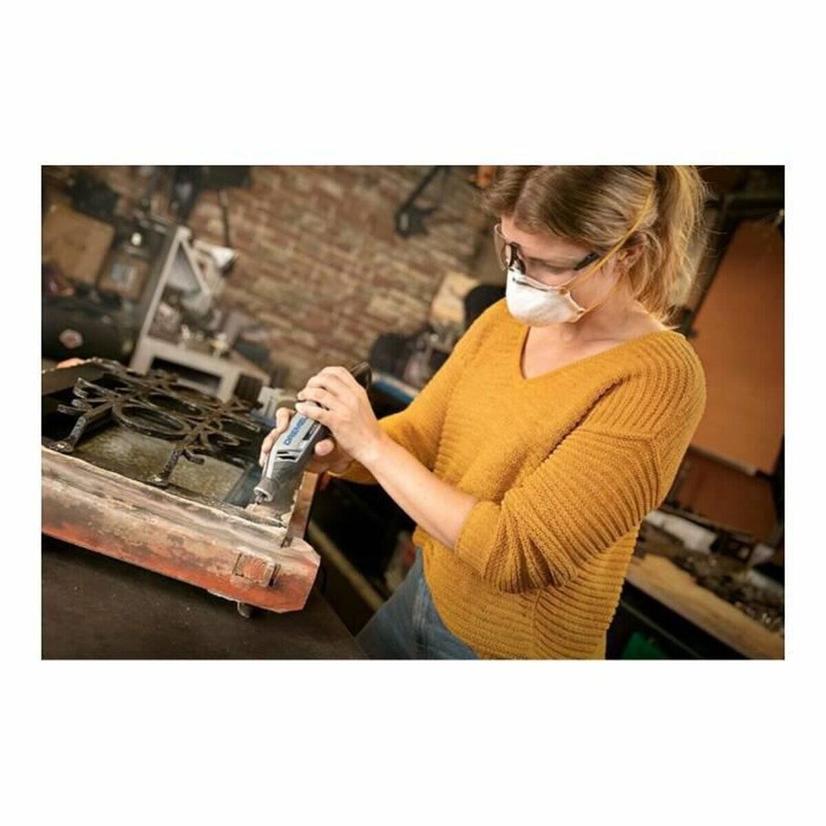 Eine Person mit Brille und Maske verwendet ein Schneidewerkzeug, um ein altes Gussmetal-Objekt in einer Werkstatt zu reparieren oder zu restaurieren.