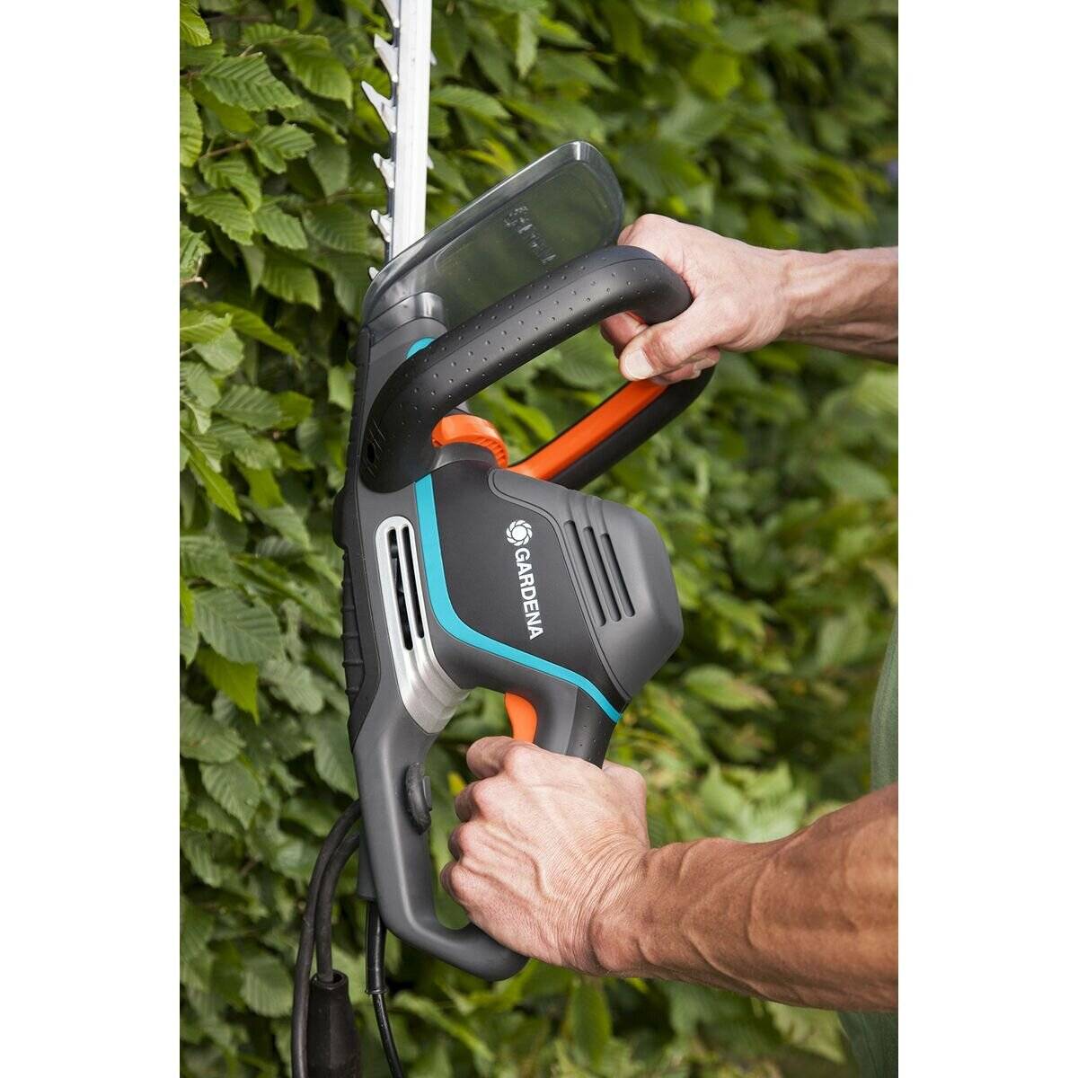 Eine Person benutzt einen elektrischen Heckenschneider, um üppiges grünes Gebüsch zu schneiden. Der Heckenschneider ist grau und blau mit einem orangefarbenen Griff.