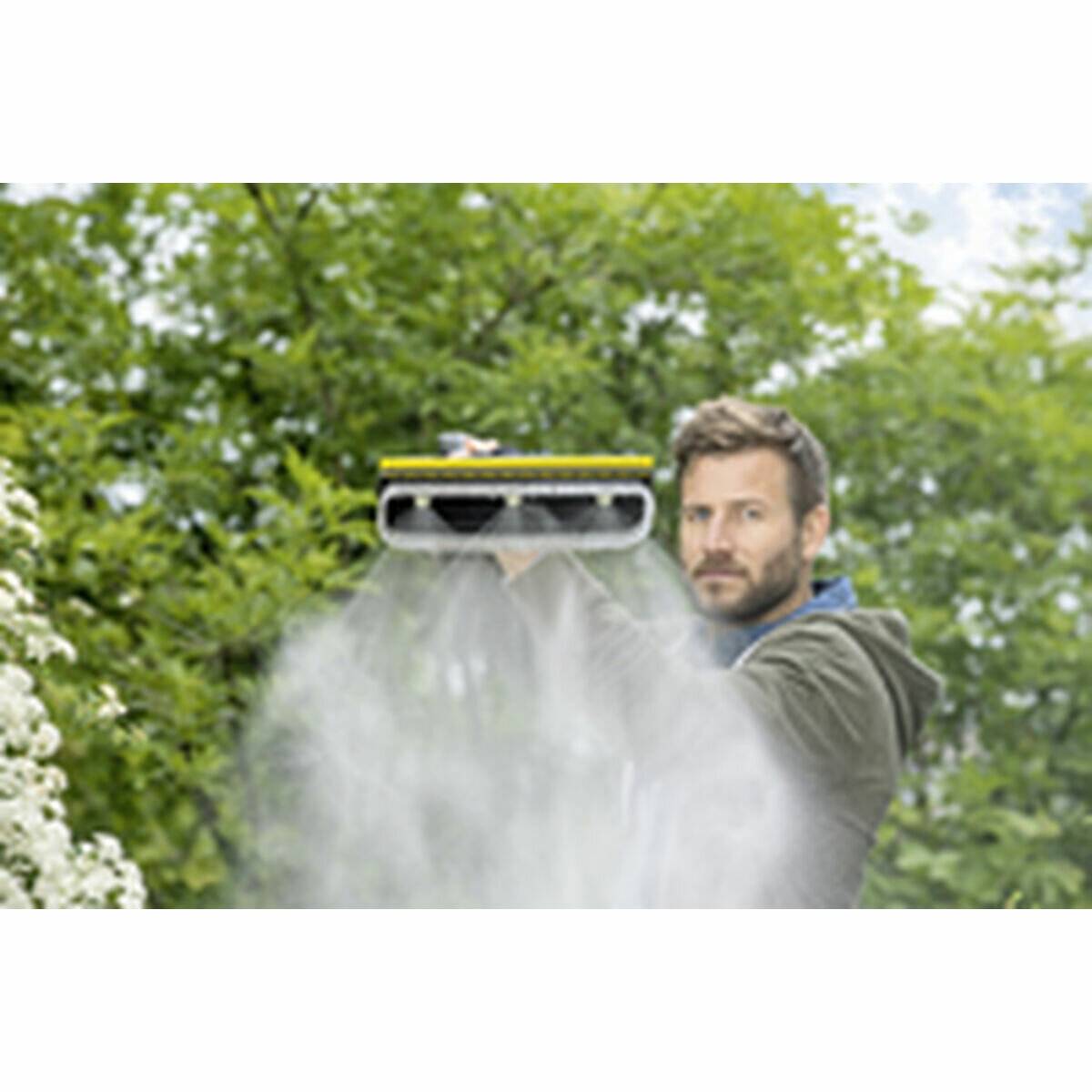 Eine Person in einer grünen Jacke verwendet einen Hochdruckreiniger auf einem Gartenweg, umgeben von Grünpflanzen, und erzeugt dabei einen nebligen Sprüheffekt.