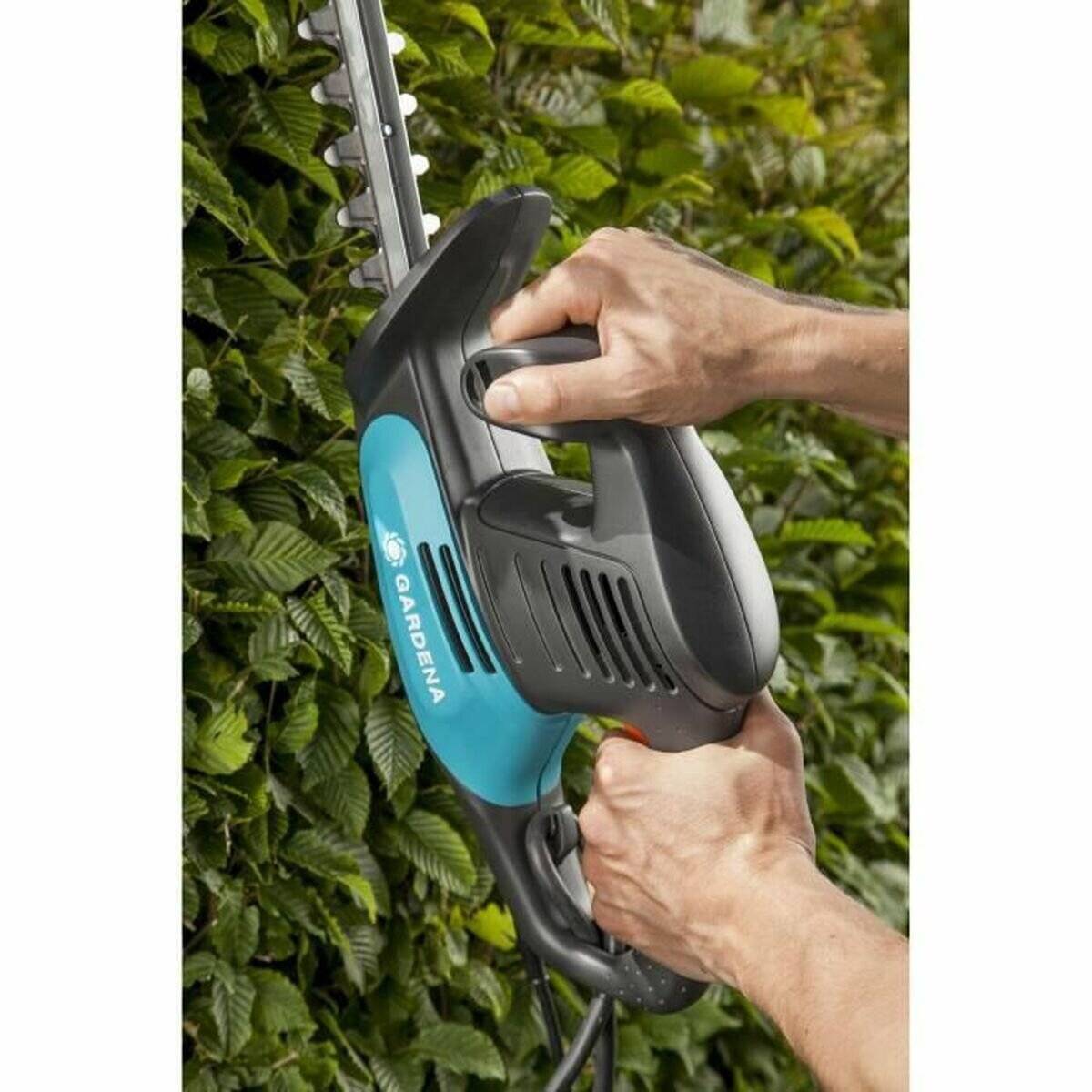 Ein Paar Hände, die einen elektrischen Heckenschneider bedienen und durch grünes Blattwerk schneiden. Das Werkzeug ist blau und schwarz, mit sichtbarem Markenschriftzug.