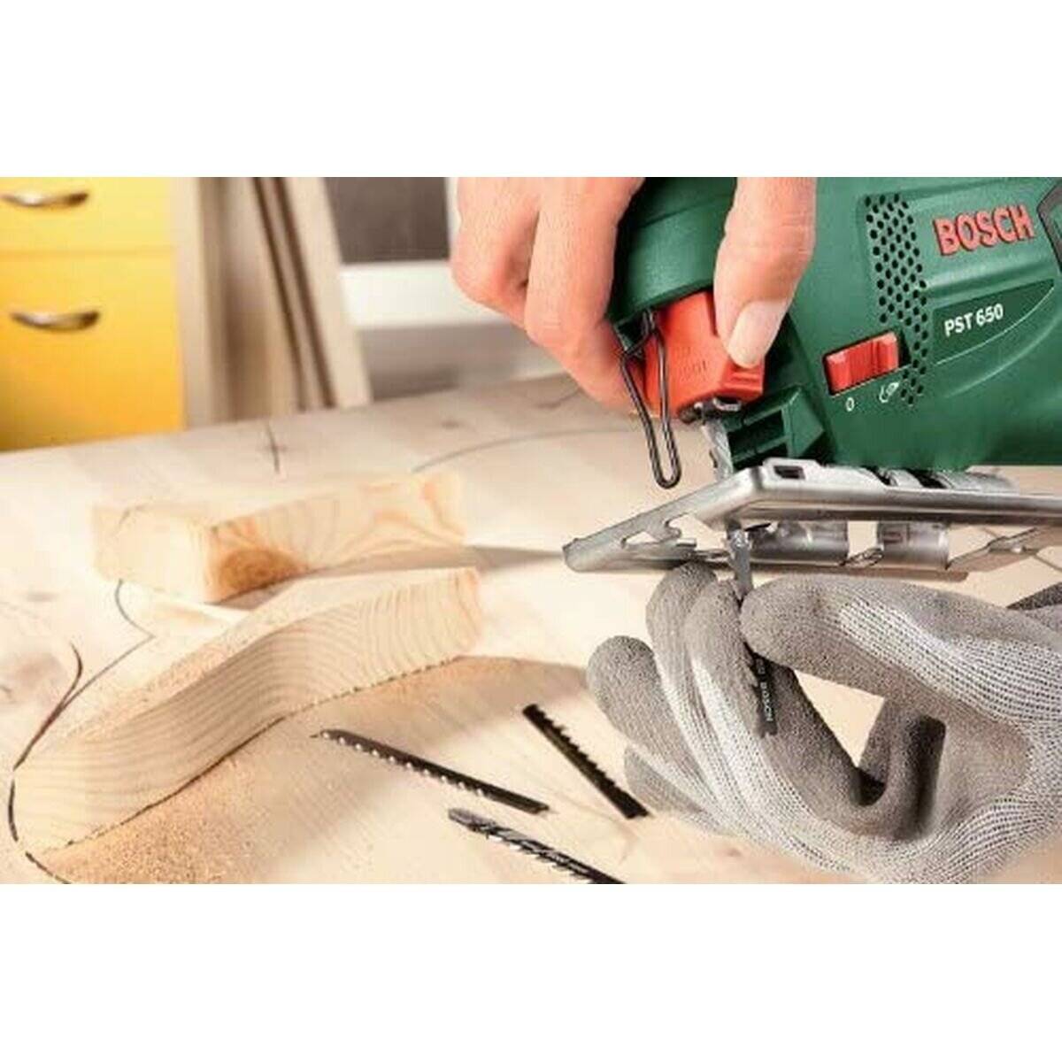 Eine Person verwendet eine Bosch PST 650 Stichsäge, um eine Holzplatte zu schneiden, mit zwei Ersatzsägeblättern und sichtbarem Sägemehl auf der Oberfläche.