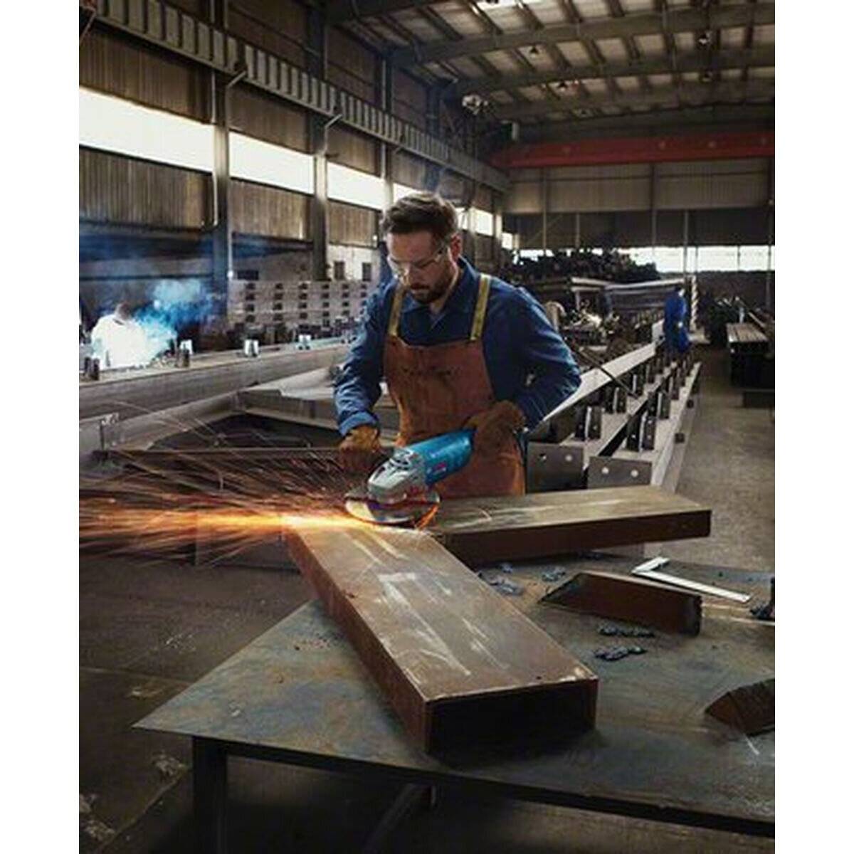 Eine Person in Schutzausrüstung schleift einen Metallträger und erzeugt dabei helle Funken in einer Industriewerkstatt mit Metallträgern und Ausrüstung im Hintergrund.