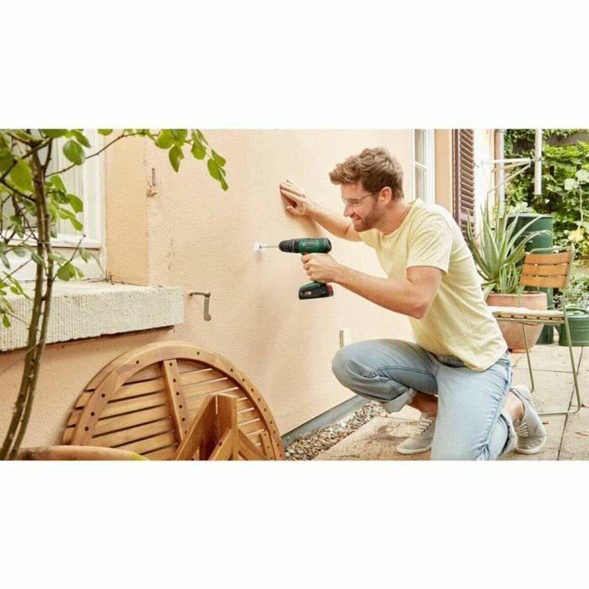 Eine Person kniet auf einer Terrasse und verwendet einen kabellosen Akkuschrauber, um eine Halterung an einer Außenwand zu befestigen. Die Umgebung umfasst Pflanzen und Gartenmöbel.