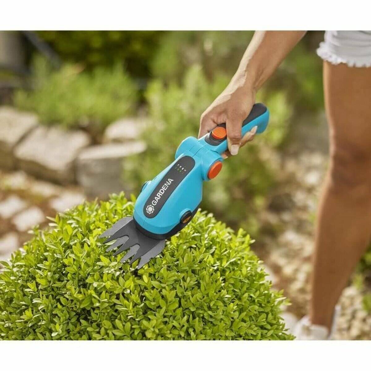 Eine Person trimmt einen Busch mit einem Gardena elektrischen Strauchschneider in einem Garten. Der Trimmer ist blau und orange. Die Szene deutet auf eine Gartenbeschäftigung hin.