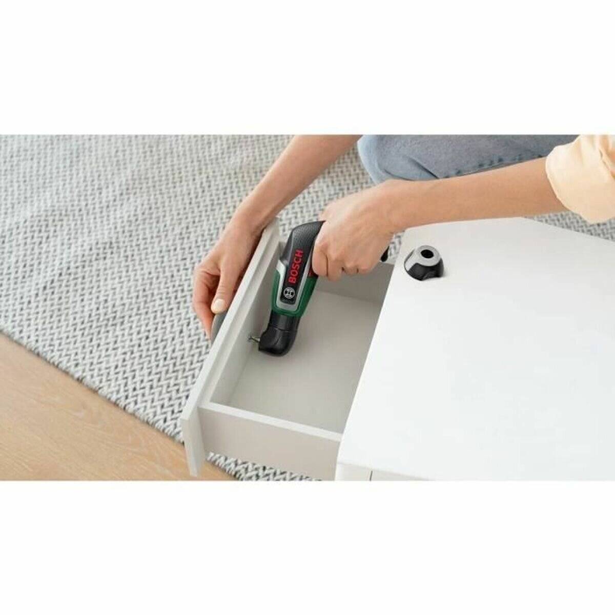 Eine Person, die einen kleinen elektrischen Schraubendreher verwendet, um ein weißes Möbelstück zusammenzubauen oder zu zerlegen, mit Fokus auf das Werkzeug und die Schublade.