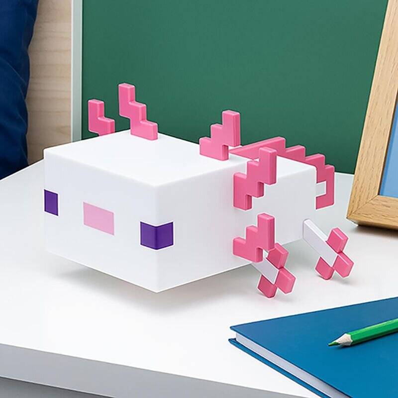 Eine Spielzeugfigur, die einem Axolotl mit eckigen Merkmalen und pinken Vorsprüngen ähnelt, steht auf einem Schreibtisch neben einem blauen Notizbuch und einem gerahmten Foto.
