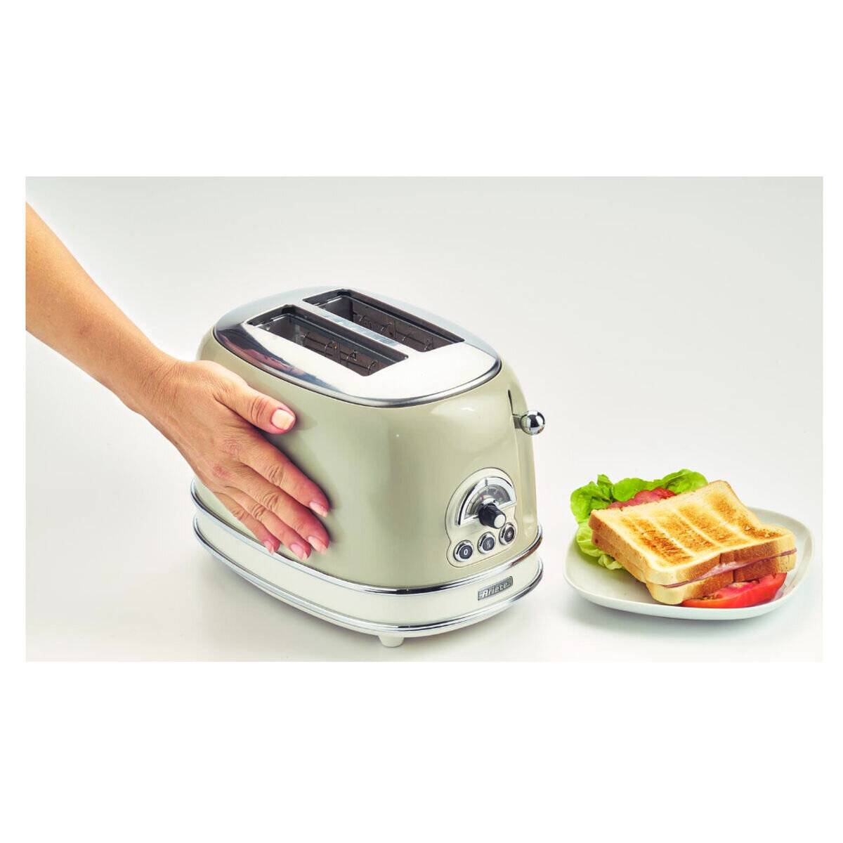 Eine Hand berührt einen Toaster im Retro-Stil mit zwei Schlitzen neben einem Teller mit getoastetem Brot und geschnittenen Tomaten auf einer weißen Oberfläche.