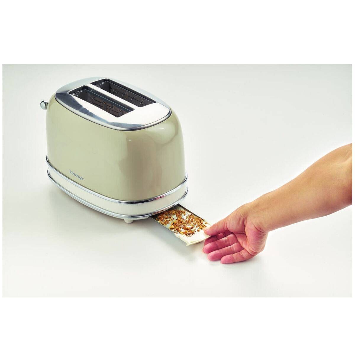 Eine Hand leert Krümel aus einem herausnehmbaren Tablett eines hellbraunen Zweischlitz-Toasters auf einer weißen Oberfläche und zeigt dabei, wie einfach die Reinigung ist.