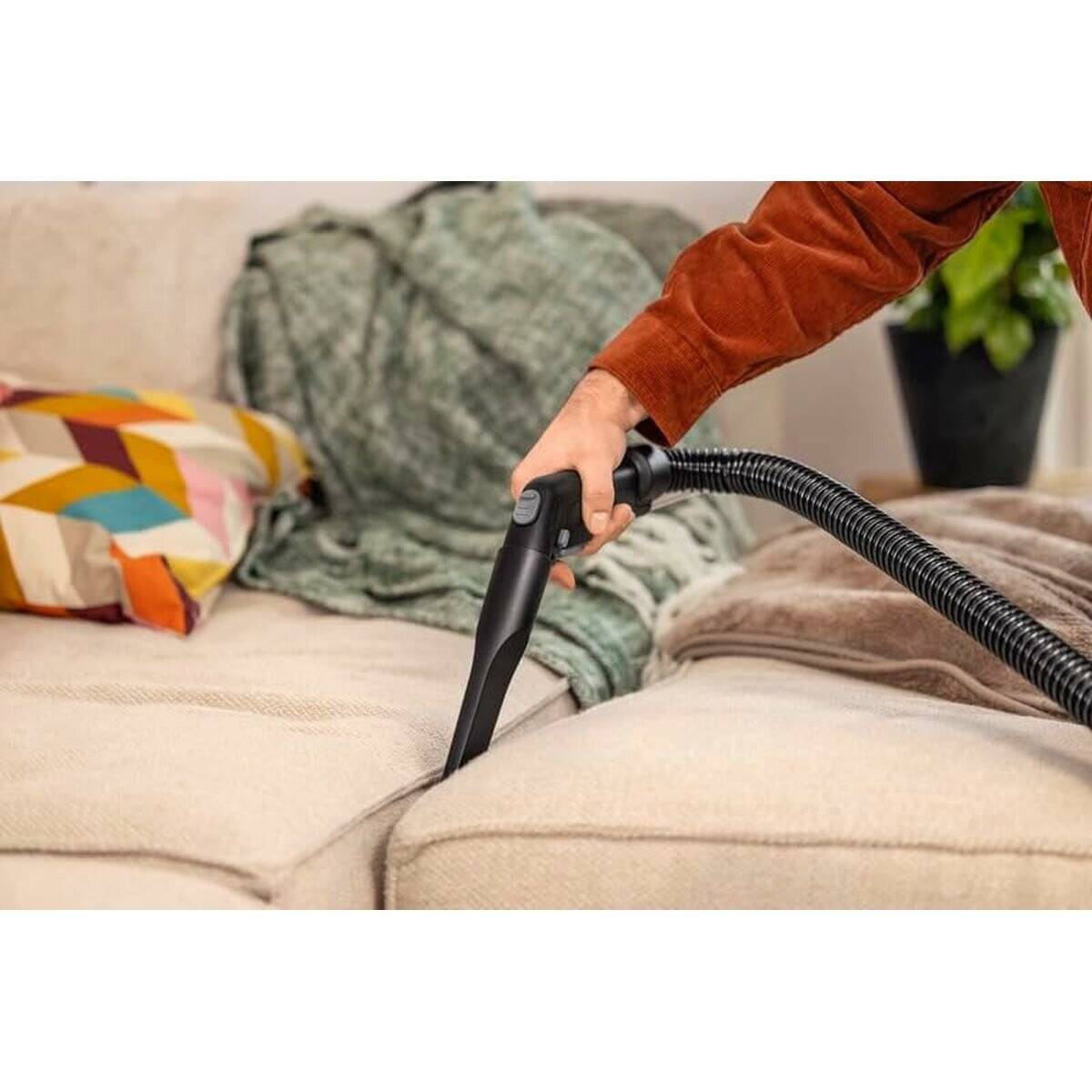 Eine Person, die einen beigen Sessel mit einem Handstaubsauger-Aufsatz reinigt, umgeben von bunten Kissen und einer grünen Decke, im Innenbereich.