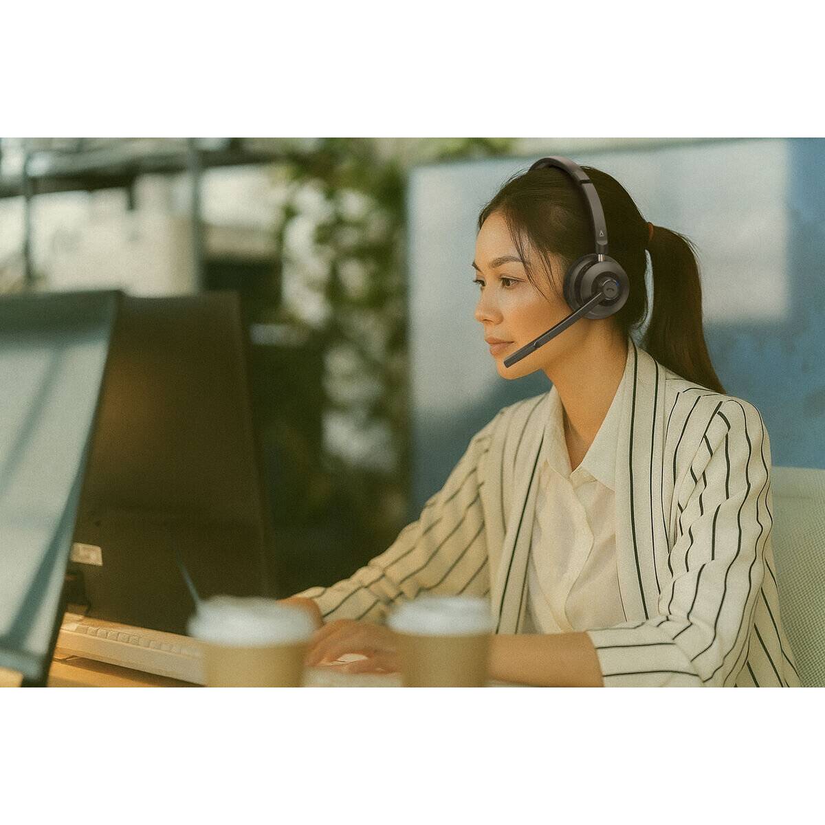 Eine Person mit gestreiftem Blazer und Headset sitzt an einem Computer und konzentriert sich auf den Bildschirm, möglicherweise an einer virtuellen Besprechung oder einem Anruf teilnehmend.
