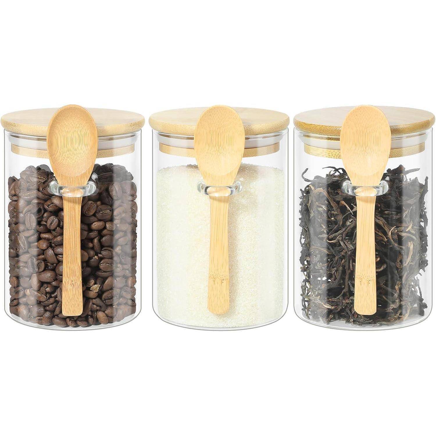Drei Glasgläser mit Holzdeckeln und Löffeln: links gefüllt mit Kaffeebohnen, Mitte mit Zucker, rechts mit getrockneten Teablättern.