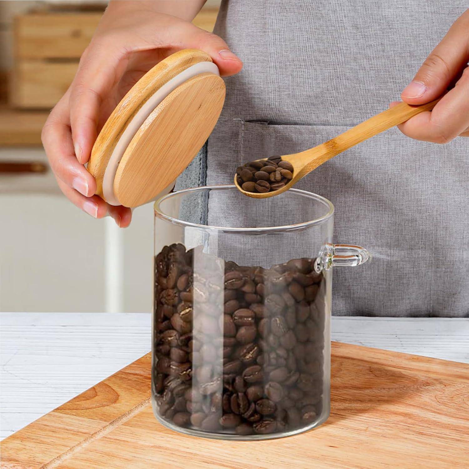 Eine Person hält einen Holzdeckel, während sie mit einem Holzlöffel Kaffeebohnen aus einem Glasgefäß schöpft, auf einer hellen Holzarbeitsplatte.
