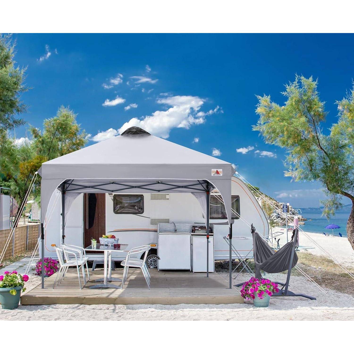 Ein Zelt mit Vordach deckt einen Essbereich mit Stühlen und einem Tisch an einem sandigen Strand ab. Bäume, Blumen und das Meer sind im Hintergrund sichtbar.