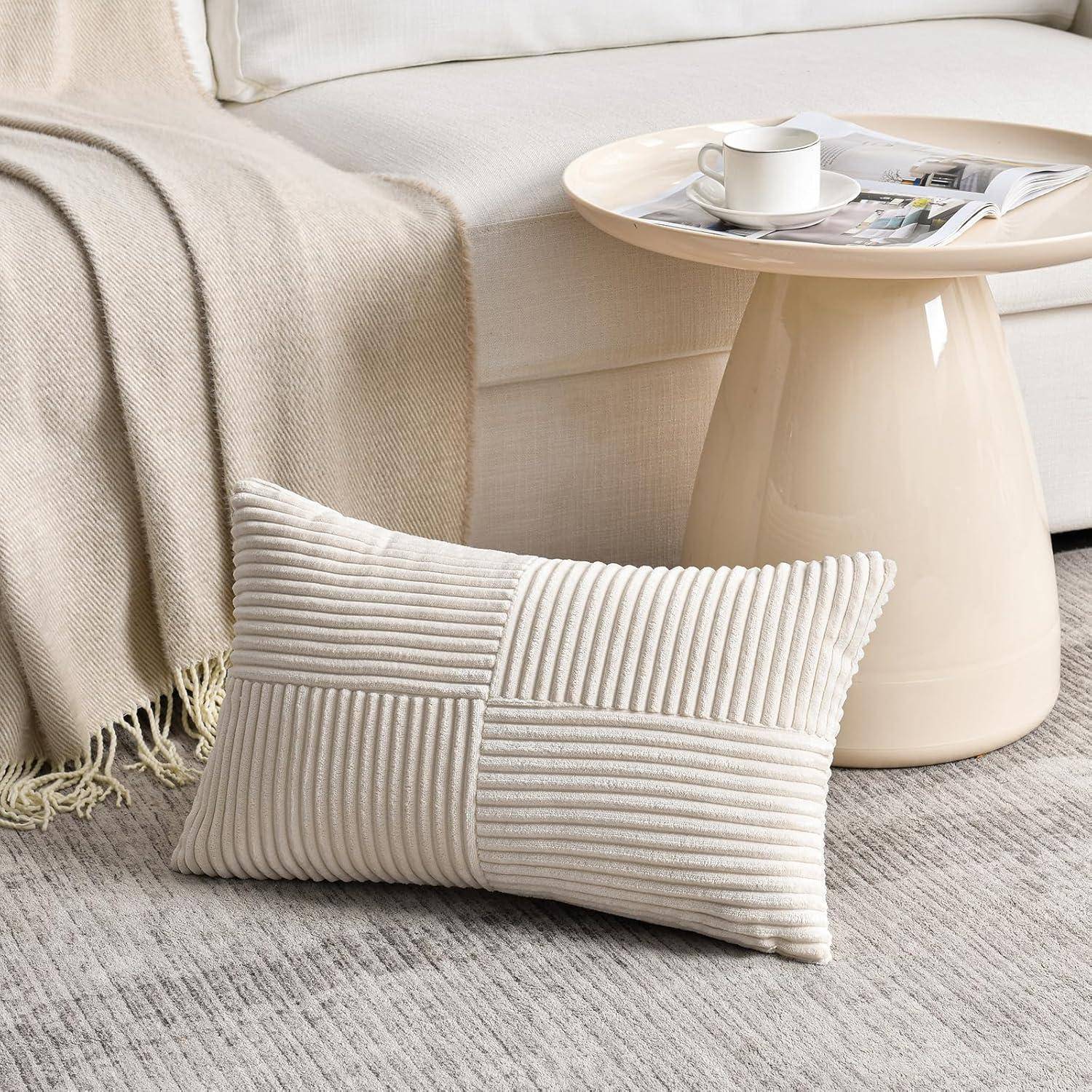 Ein hellbeiger Sofa mit einer zusammengelegten Decke und einem strukturierten Kissen. Ein kleiner runder Tisch trägt eine Zeitschrift und eine weiße Tasse neben dem Sofa.