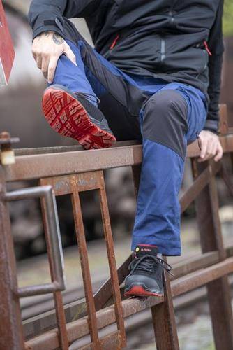 Eine Person trägt blaue Hosen und Turnschuhe mit leuchtend roten Sohlen und tritt auf eine Metalloberfläche, wobei die Traktion und der Stil des Schuhs hervorgehoben werden.