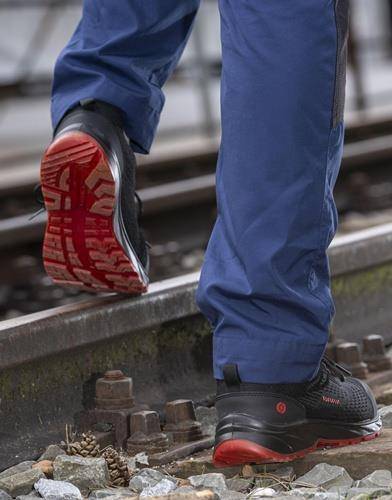 Eine Person mit blauen Hosen und schwarz-roten Wanderschuhen überschreitet Gleise und konzentriert sich dabei auf das Profil und das robuste Design der Schuhe.