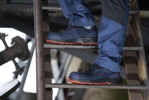 „Person mit blauen Arbeitsschuhen und roten Sohlen, die auf einer Metalltreppe steht.