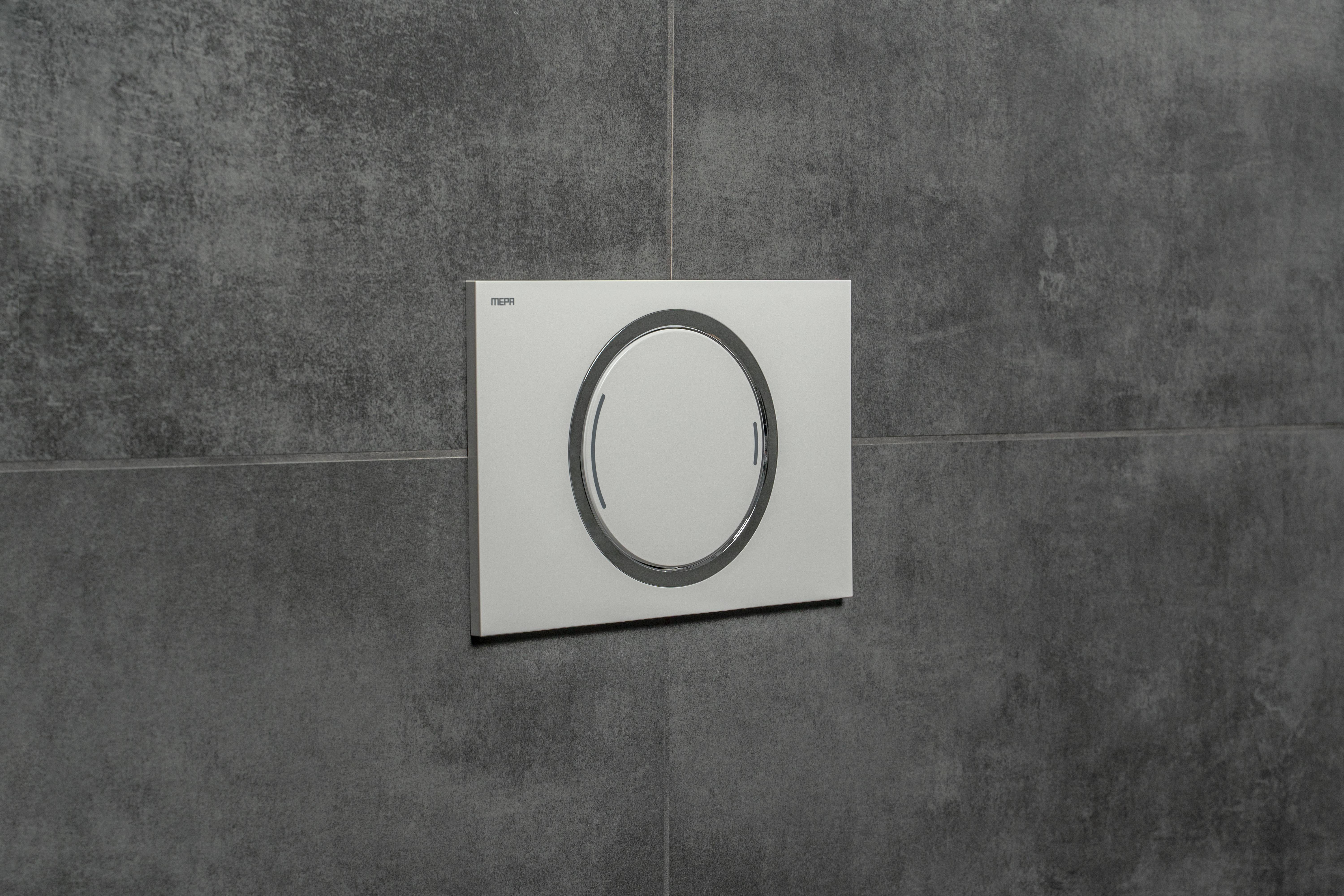 Eine weiße, kreisförmige Betätigungsplatte, die an einer grauen gefliesten Wand montiert ist.