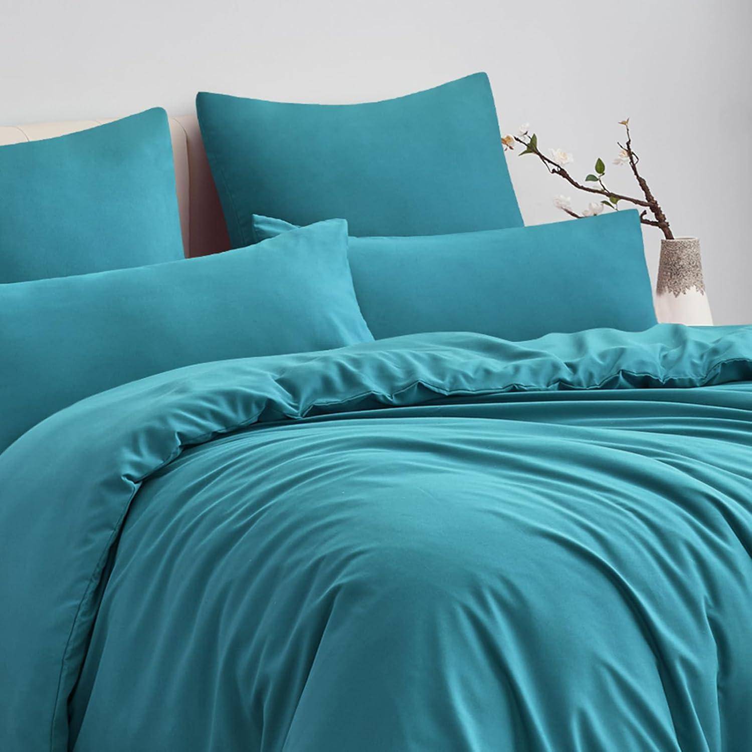 Ein ordentlich gemachtes Bett mit Petrol-Bettwäsche, bestehend aus Kissen und Bettdecke, neben einer Vase mit einem kleinen Zweig vor einem minimalistischen Hintergrund.