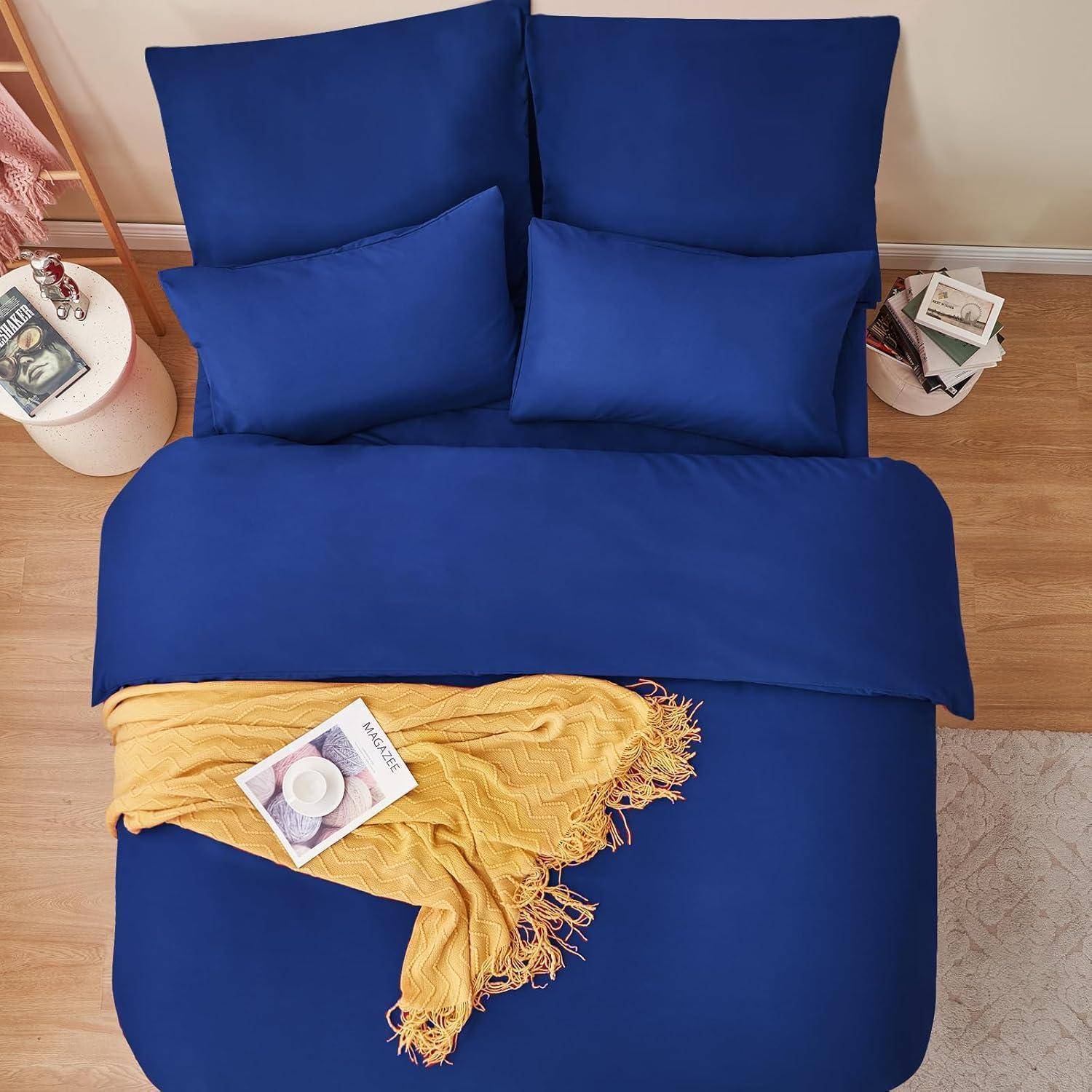 Ein ordentlich hergerichtetes Bett mit lebhafter blauer Bettwäsche, zwei großen und zwei kleinen Kissen, einer gelben Decke und einer Zeitschrift. Ein Holzboden und ein kleiner Beistelltisch mit Büchern und einer Pflanze sind zu sehen.